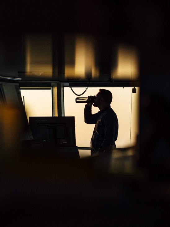 A silhouetted deck officer drinks from a mug while looking out illuminated windows on the bridge of icebreaker Kontio.