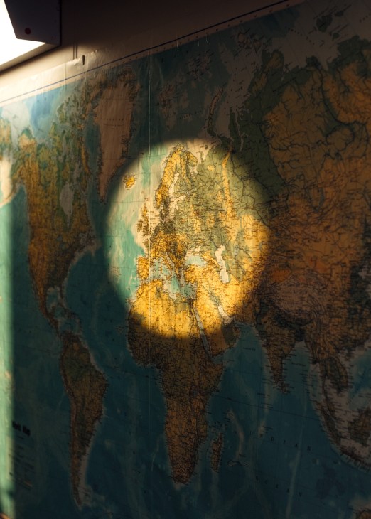An antique world map illuminated by sunlight on a ship's wall, showing continents in gold and green tones.
