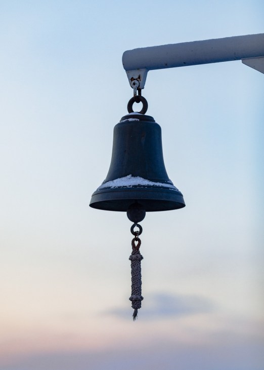A ship's bell with rope lanyard hanging from a metal bracket against a clear sky.