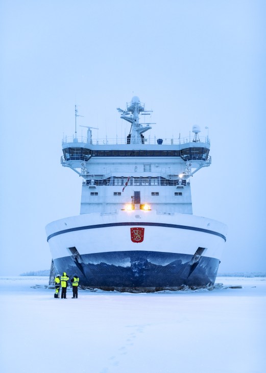 Finnish icebreaker IB Kontio docked in frozen Bay of Bothnia with three crew members in yellow safety gear standing on ice below.