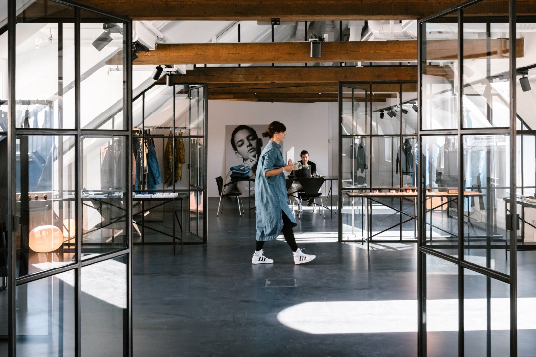 Woman in denim shirt pacing through Tom Wood's minimalist loft space with exposed wooden beams and floor-to-ceiling windows.