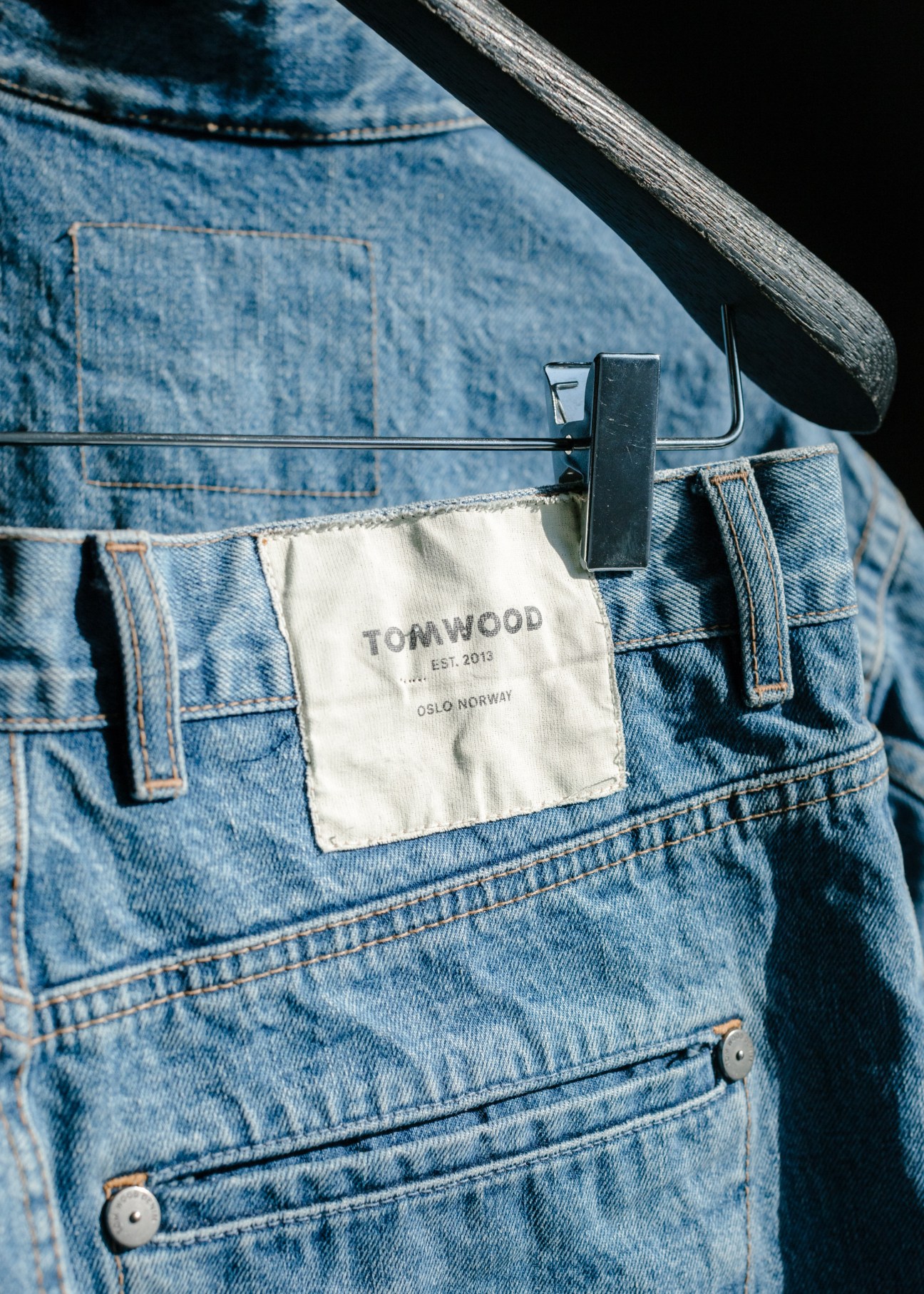 Close-up of Tom Wood denim with cream label tag clipped to waistband against wooden railing.