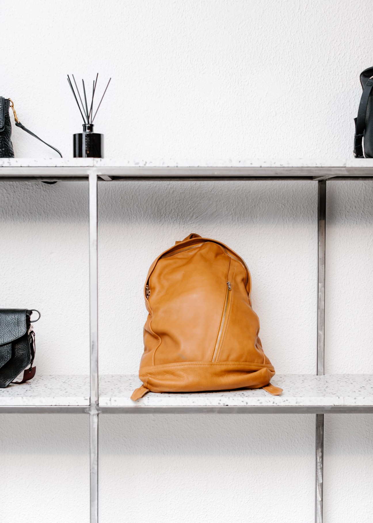 Orange leather backpack displayed on white shelving unit with black accessories and a diffuser.