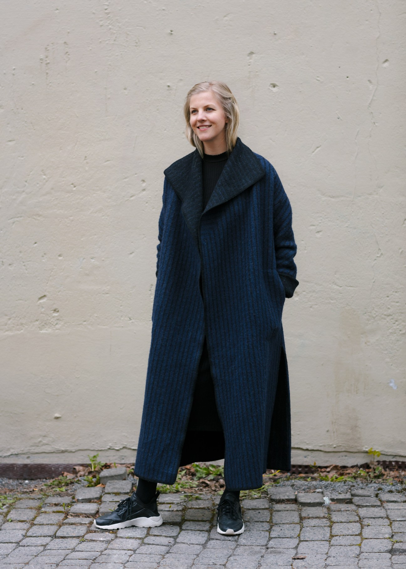 Elisabeth Stray Pedersen wears a long dark blue ribbed wool coat over black clothing against a white brick wall.