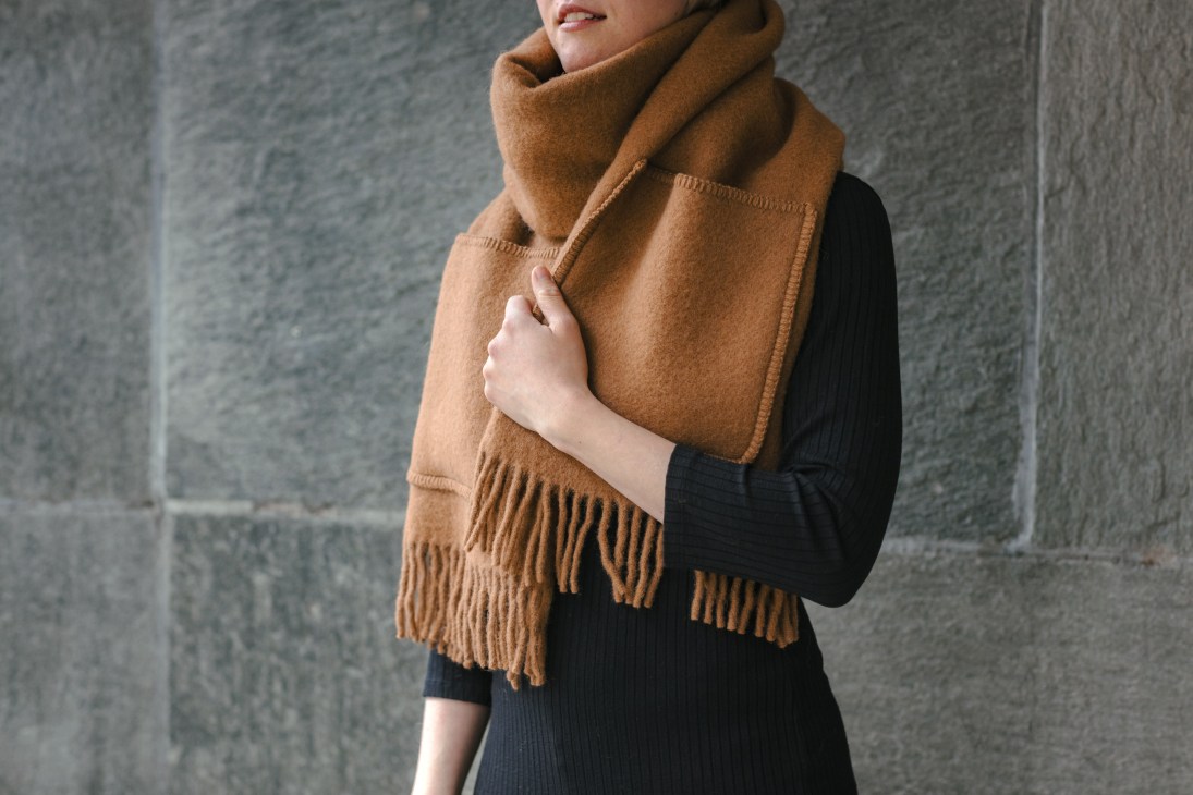 Person wearing a camel-colored wool scarf with fringe over a black ribbed sweater.