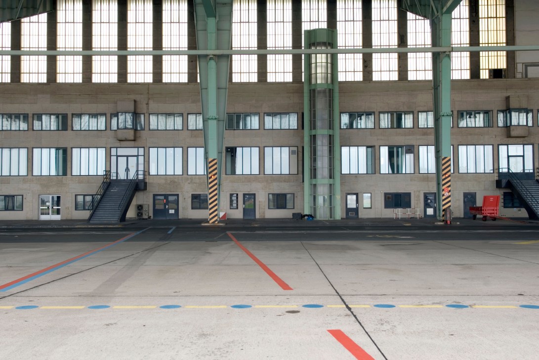 Interior of Tempelhof airport hangar with large windows, structural columns, and colored floor markings.
