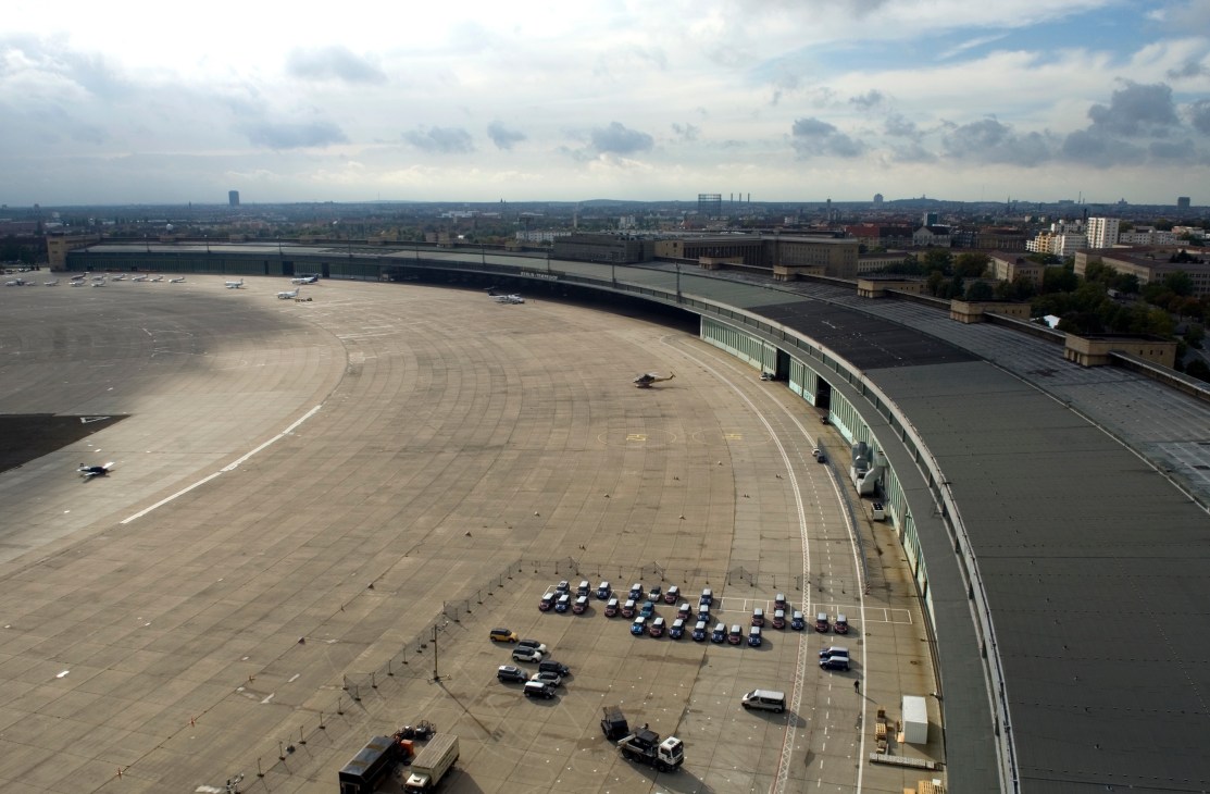 Aerial view of Tempelhof airport's vast curved tarmac with parked vehicles and Berlin cityscape beyond.