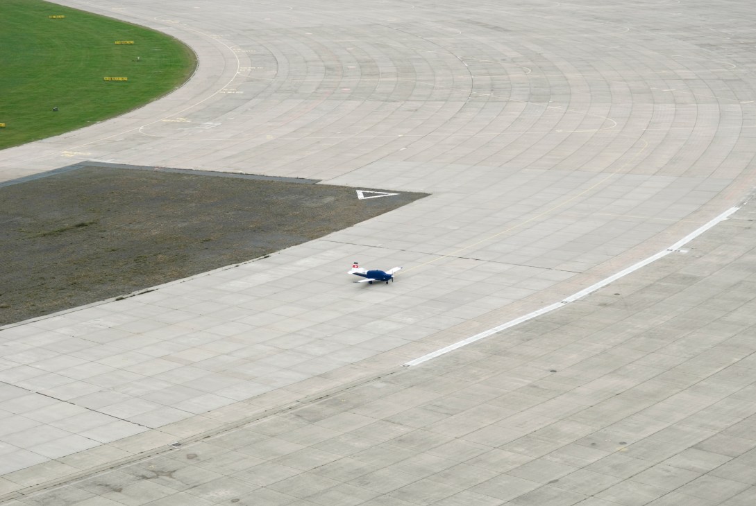 A small private jet parked on the expansive tarmac of Tempelhof airport with grass and runway markings visible.