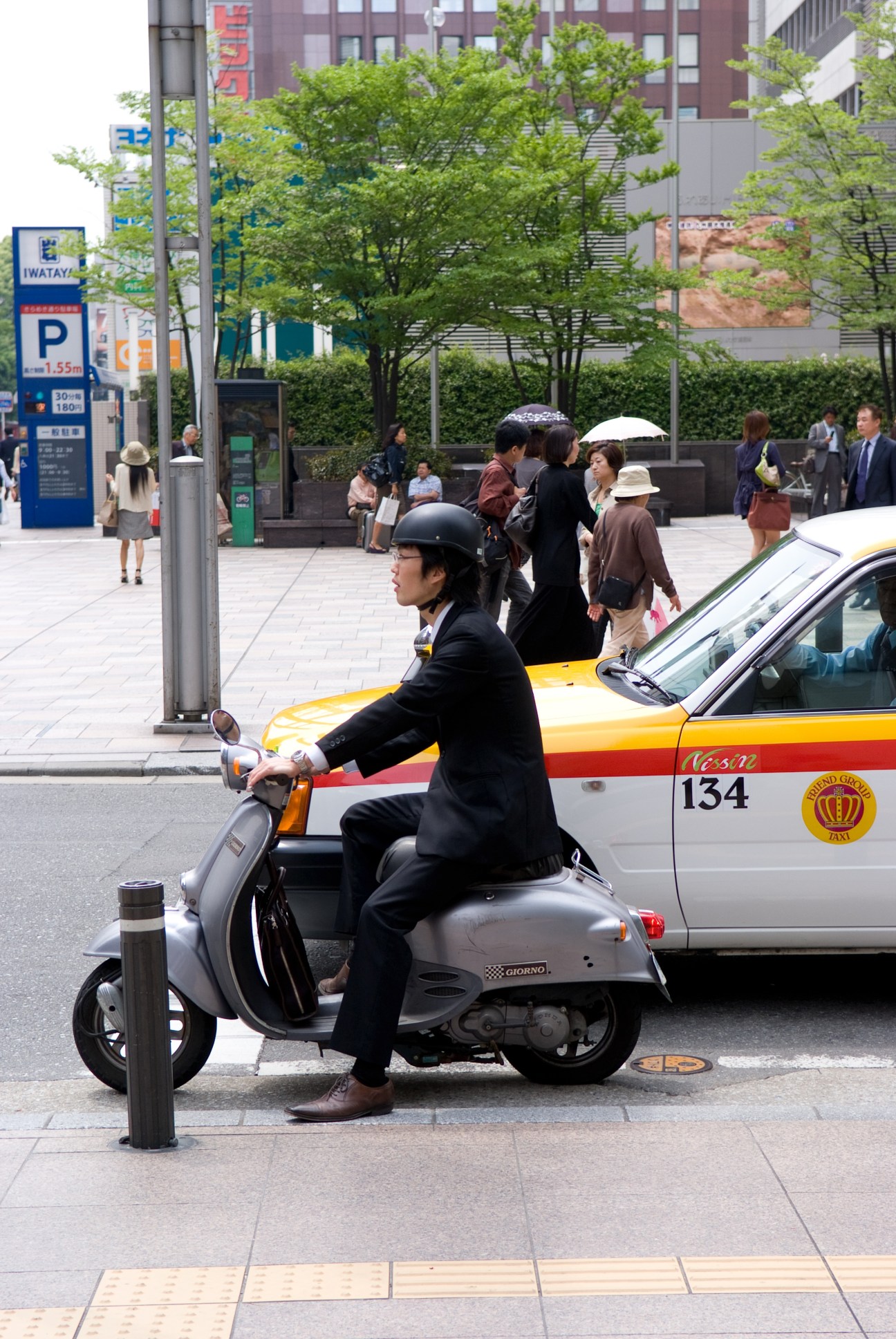 Taxi driver on a scooter in Tenjin district with pedestrians and urban buildings in background.