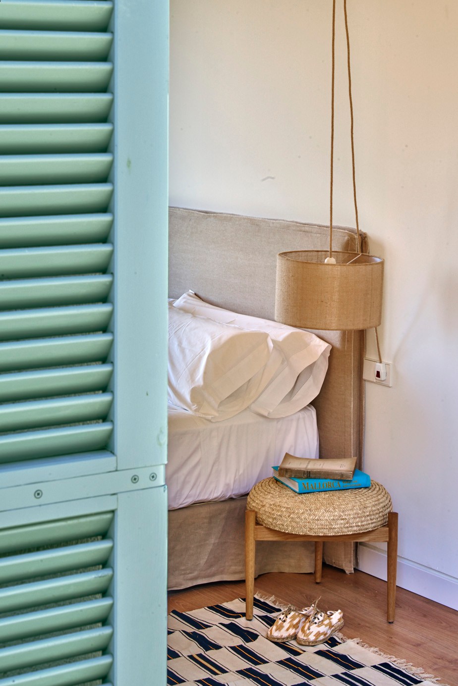 Bedroom with unmade white bed, wooden chair with woven seat, pendant lamp, books and wicker basket