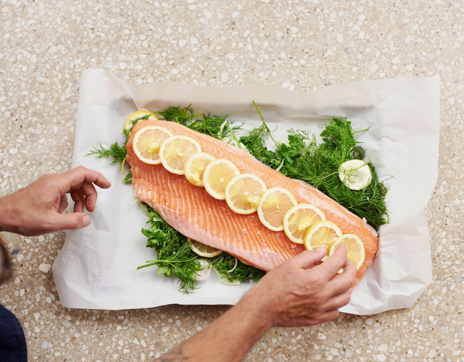 Hands holding a white plate with salmon fillet topped with lemon slices on fresh herbs.