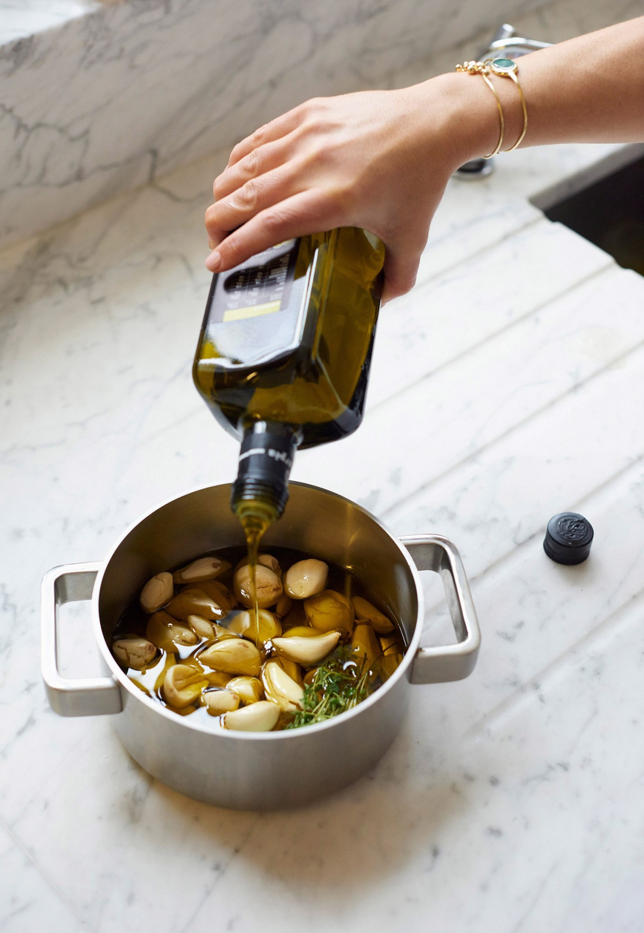 Person pouring olive oil over garlic cloves and herbs in a white pot on a marble countertop.