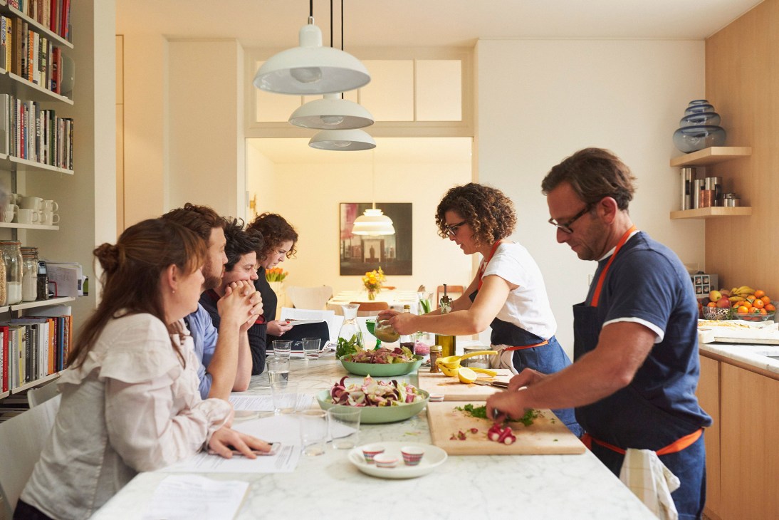 Hanna Geller Goldsmith and Jeremy Coleman teaching a cooking class to six participants in a bright kitchen with bookshelves and pendant lights.