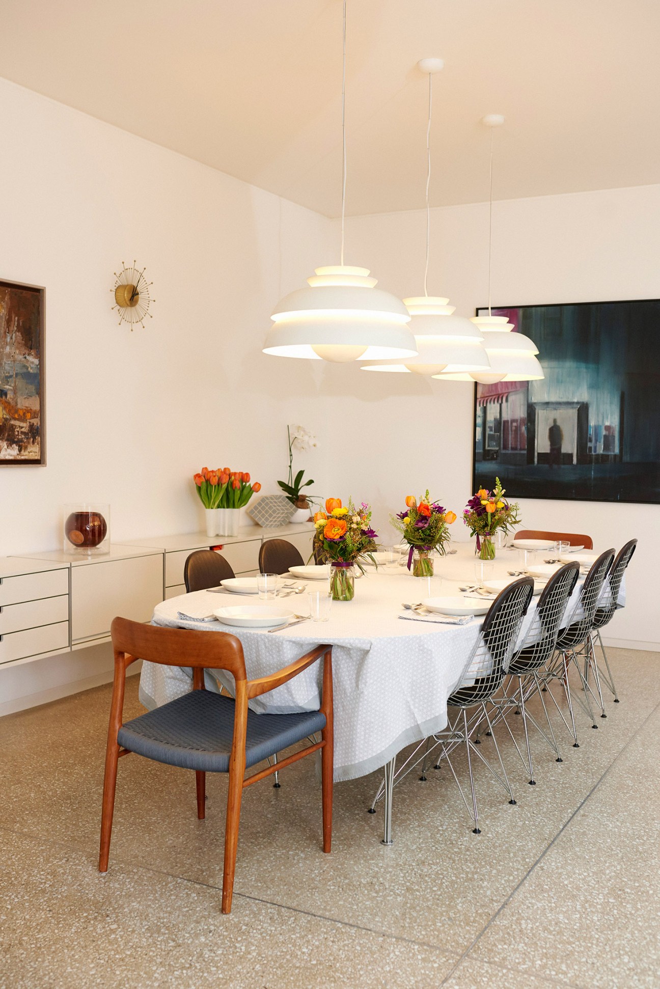 Modern dining room with round white table set for eight, pendant lights overhead, and mixed mid-century chairs.