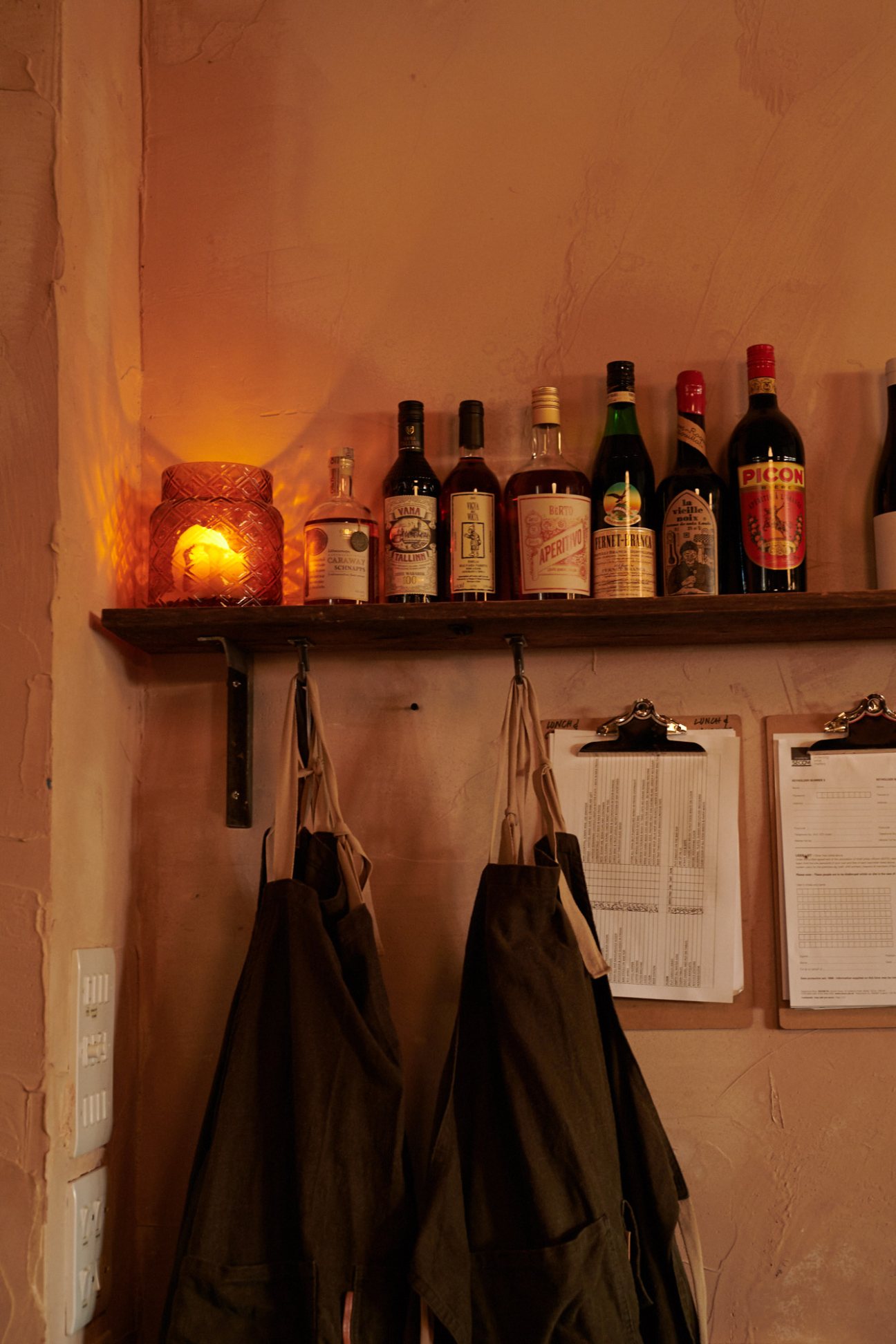 Shelf displaying bottles of wine and spirits next to an illuminated candle and hanging aprons.