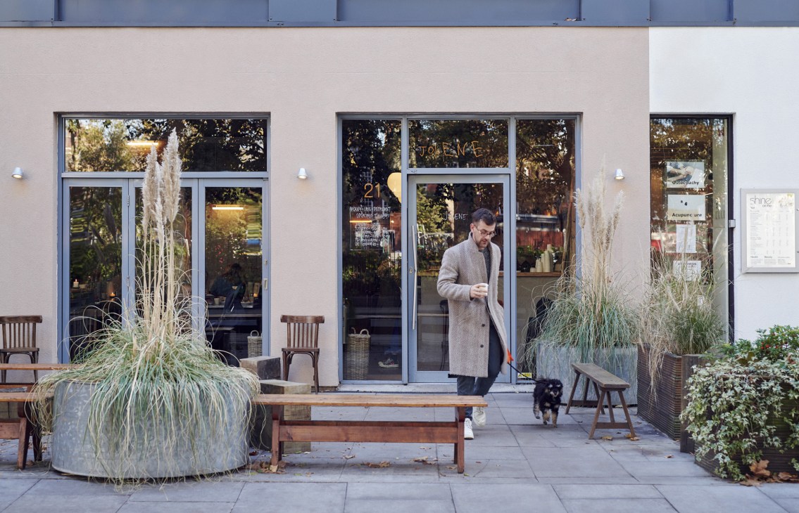 Man in beige coat standing outside Jolene restaurant on Newington Green with planted grasses and wooden chairs on the pavement.