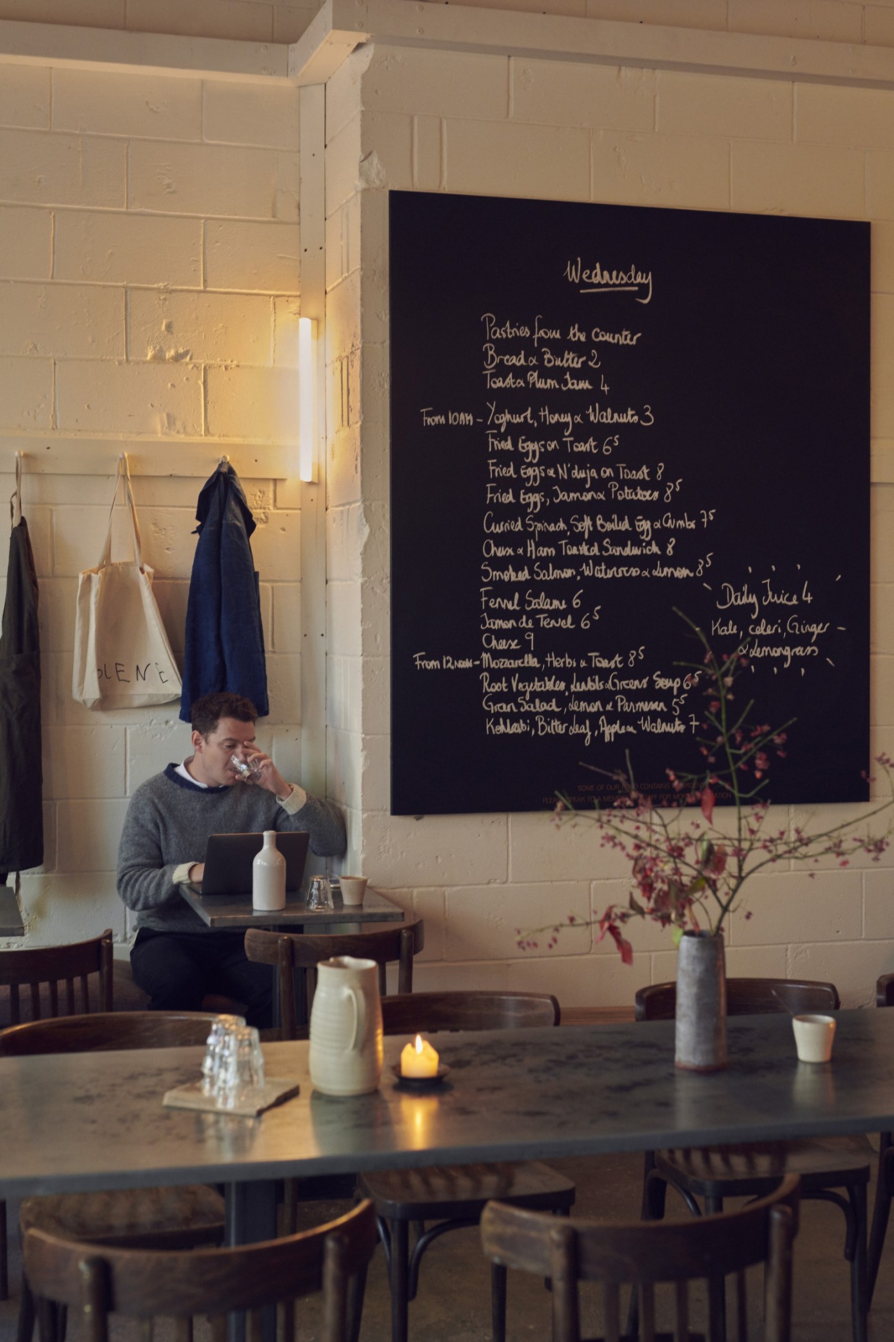 A person sitting at a small table holds a cup while looking at a laptop, beneath a black chalkboard menu displaying Wednesday's dishes at Jolene restaurant.