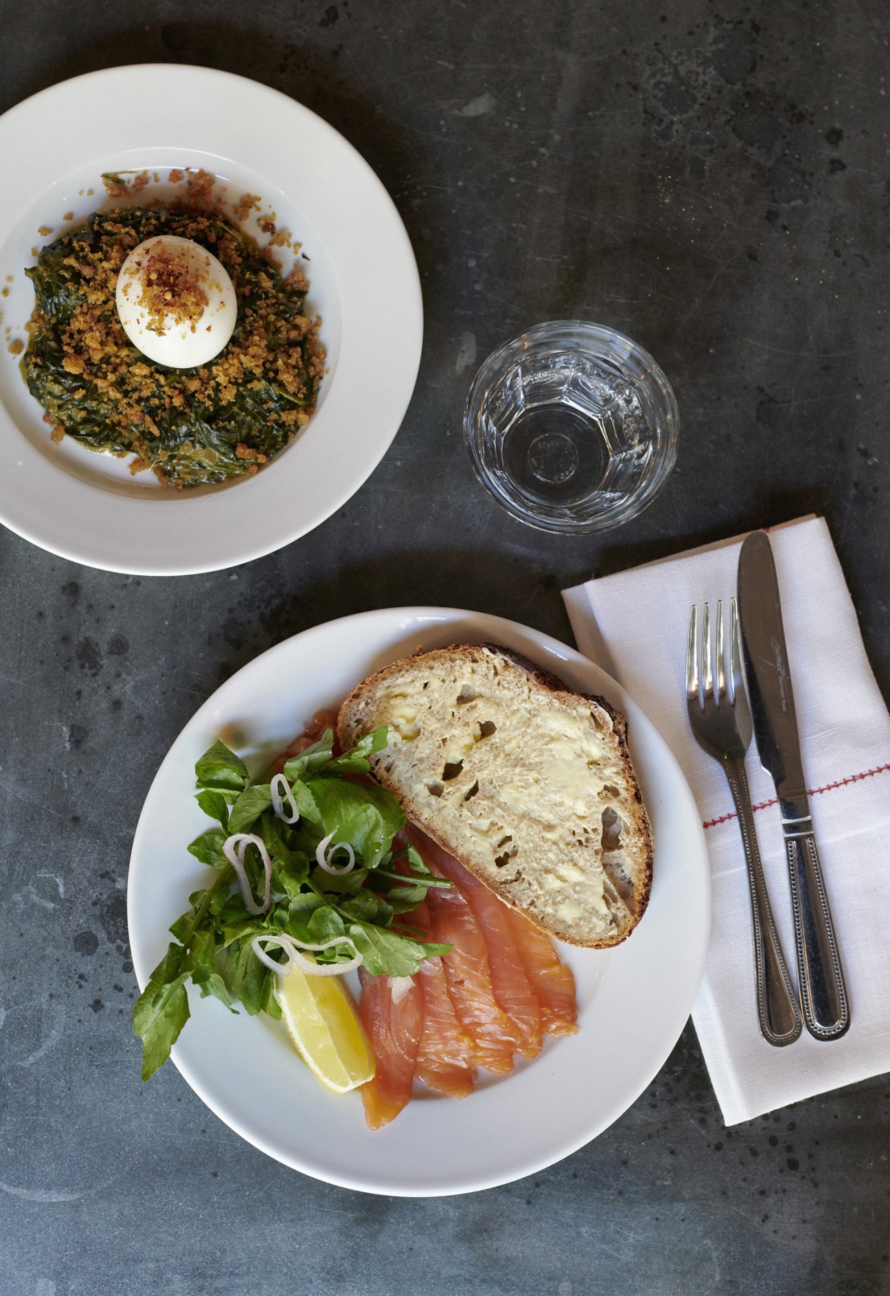 Two plates of food with smoked salmon, greens, and bread on a dark table with cutlery and glass