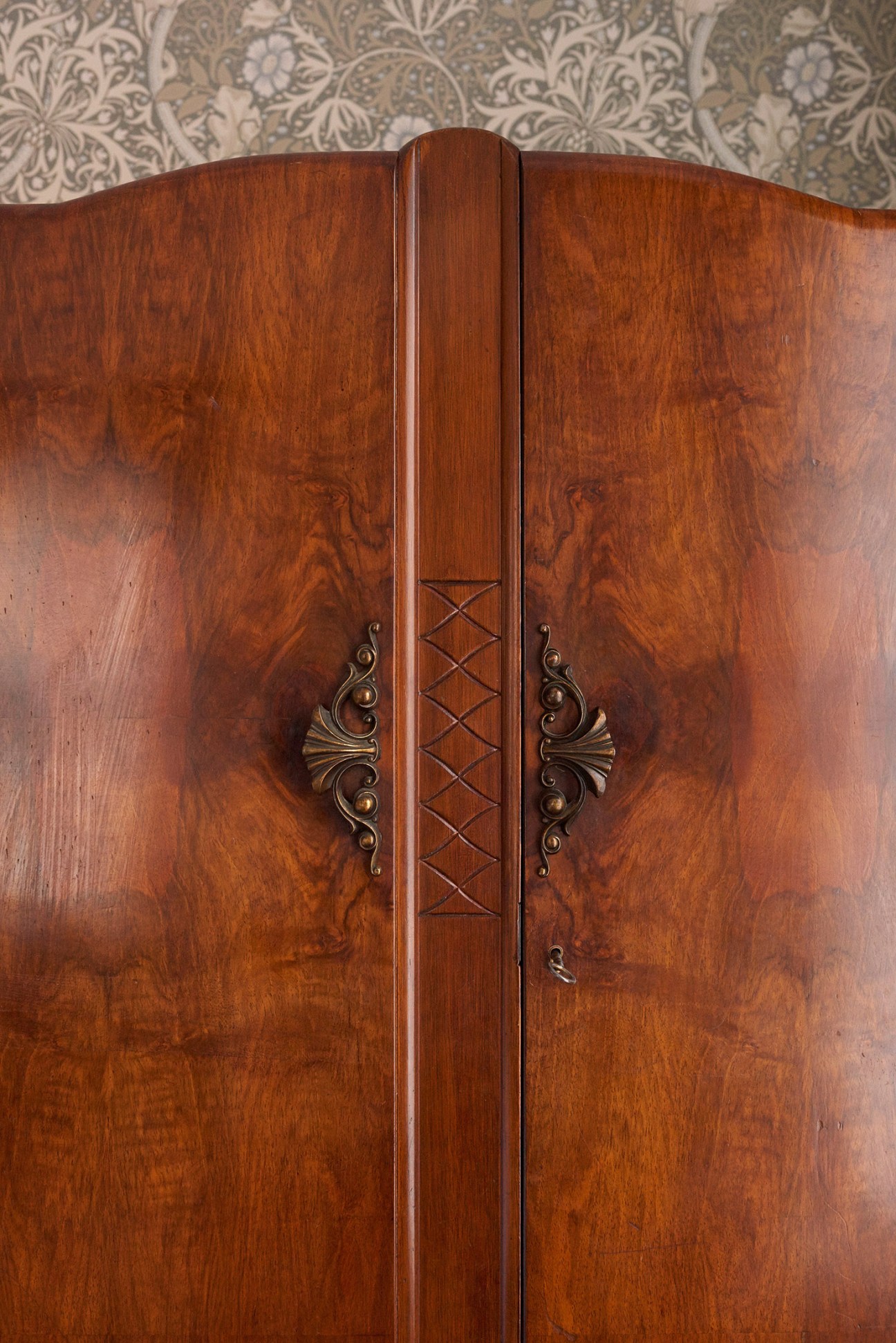 Art-deco armoire with wooden doors and ornamental brass handles against William Morris wallpaper.