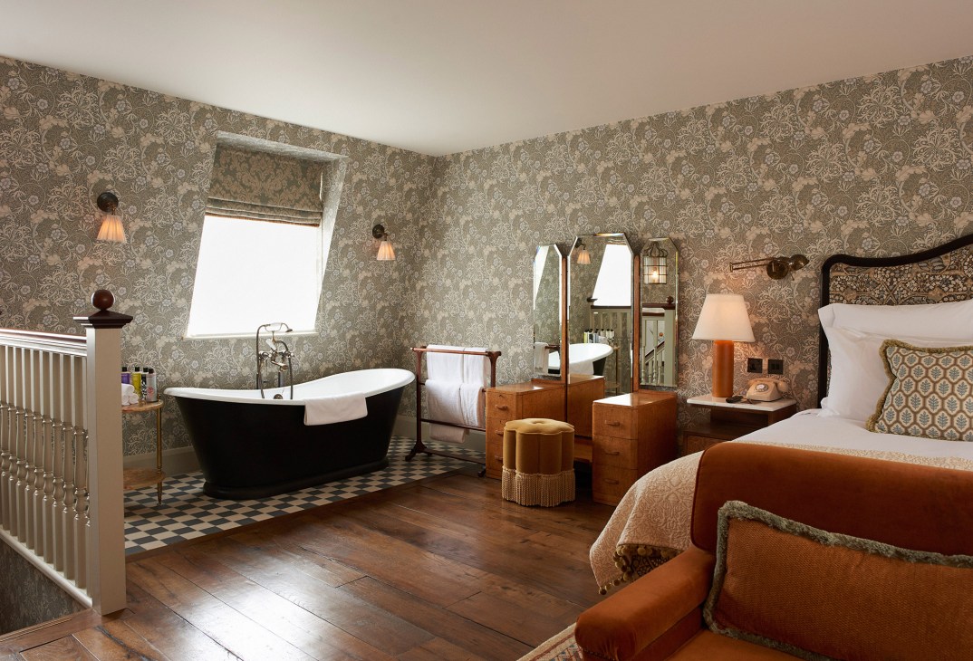 A luxurious bedroom with William Morris floral wallpaper, featuring a black freestanding bathtub, wooden flooring, and period furnishings.