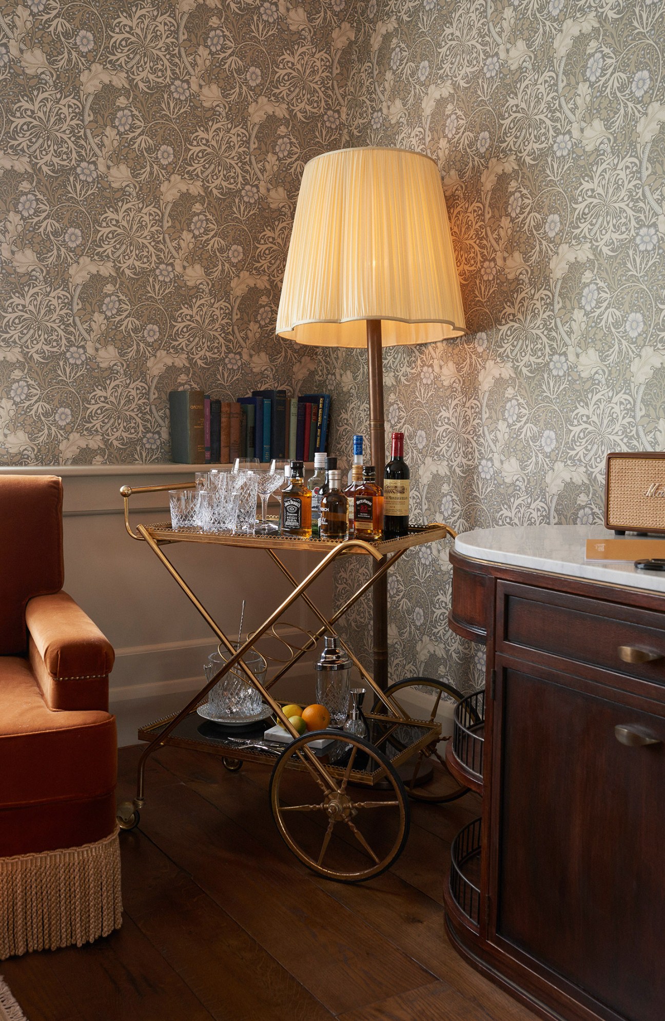 A brass drinks trolley with liquor bottles and glasses beneath a pleated cream lampshade against William Morris wallpaper.