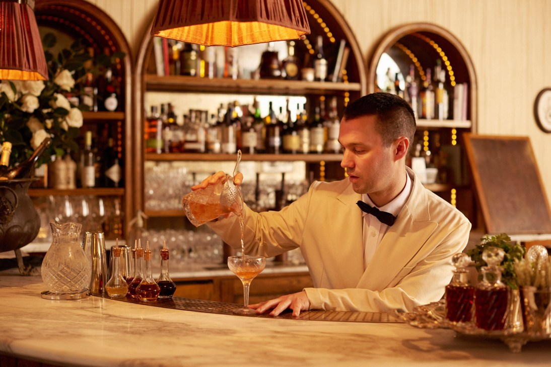 Bartender in cream shirt pouring cocktail at Kettner's bar with arched shelving and hanging lights behind.