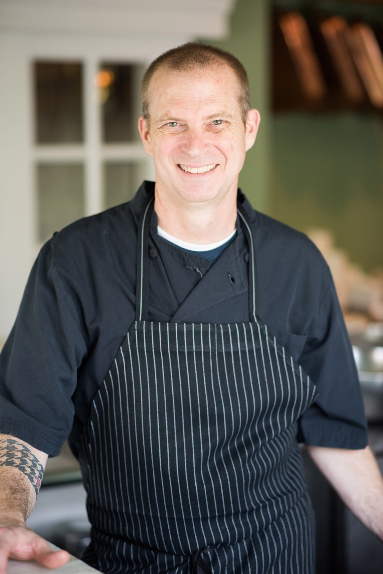 Chef Matthew Bousquet smiles at The Clifton restaurant wearing a black pinstriped apron.