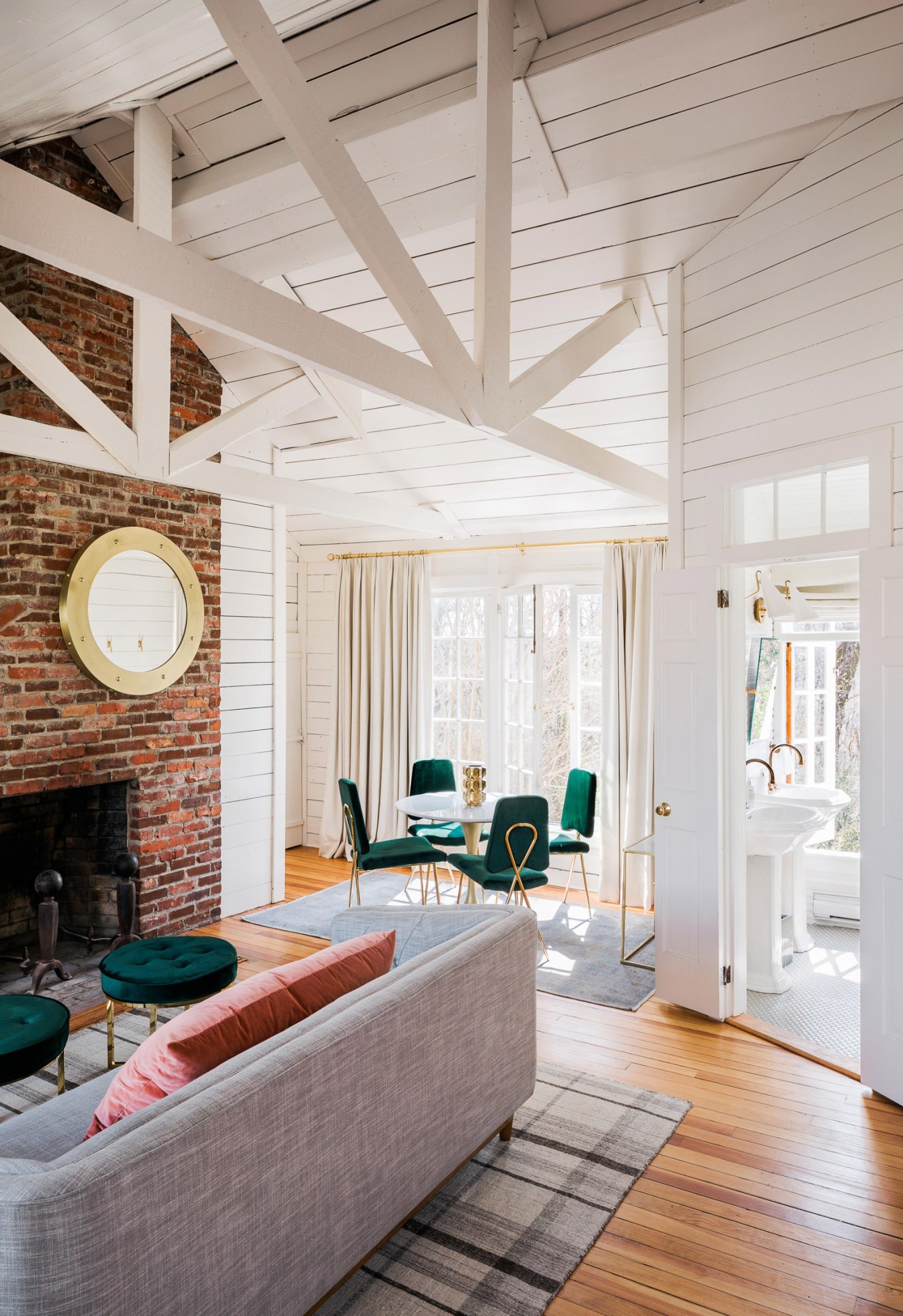 Bright modern interior with exposed brick fireplace, white beamed ceiling, gray sofa, green velvet chairs, and cream curtains at The Clifton bridal suite in Virginia.