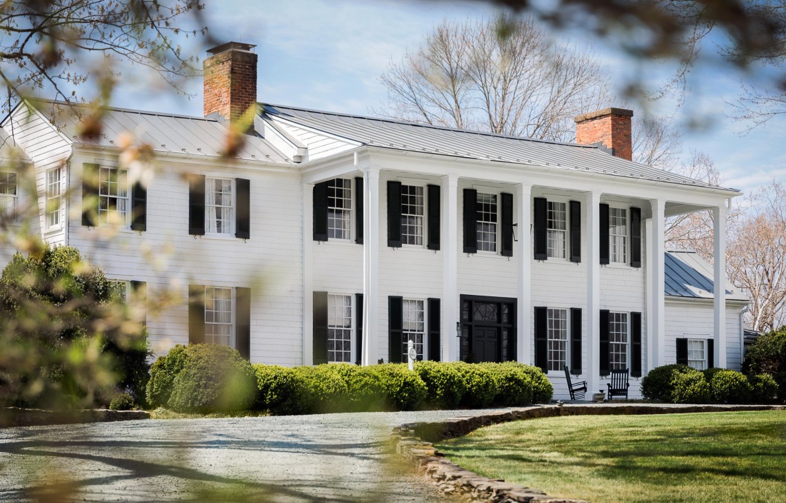 White colonial-style manor house with black shutters, brick chimneys, columned porch, and metal roof.