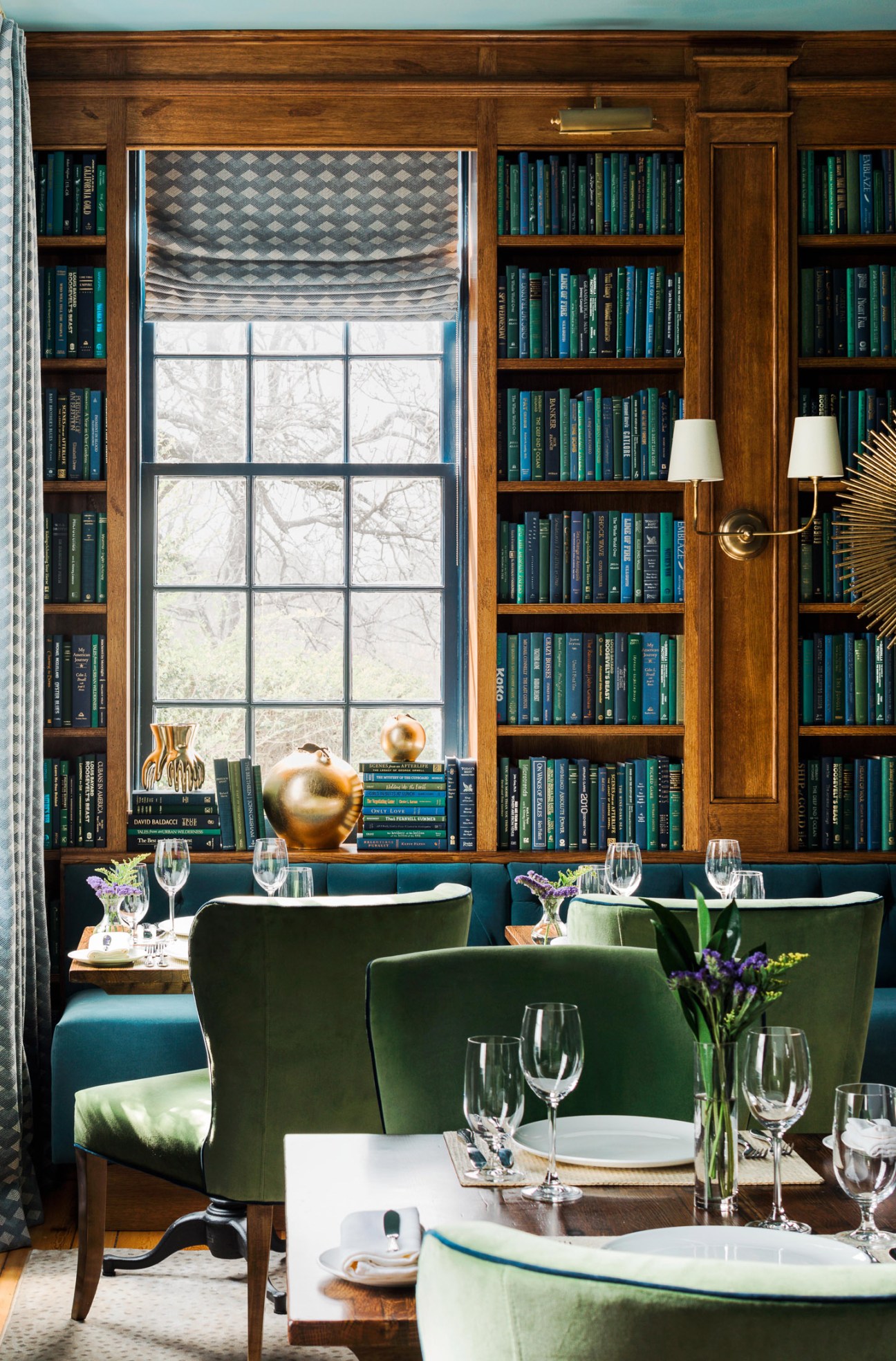 Elegant restaurant dining room with wooden bookshelves, green velvet chairs, and large window overlooking bare trees.