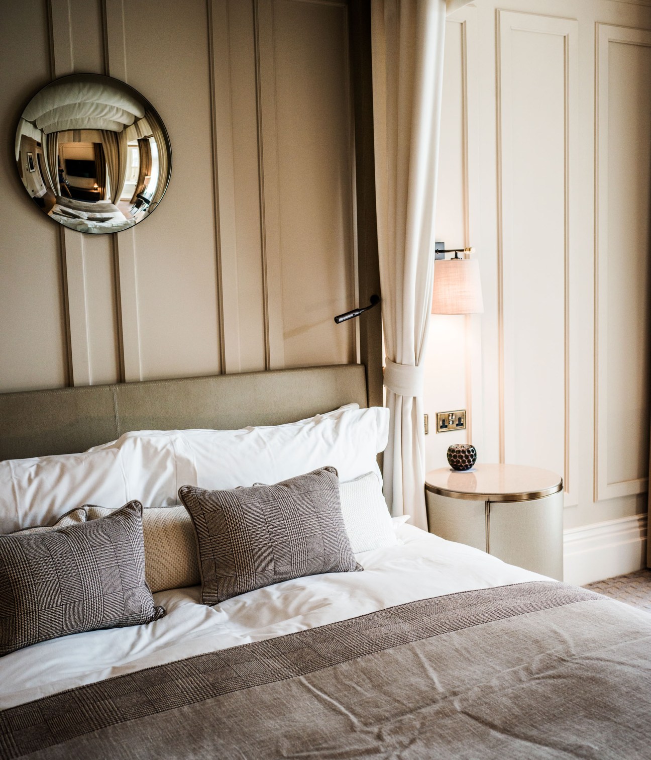 Modern hotel bedroom with neutral tones, patterned pillows, circular mirror, and cream panelled walls.