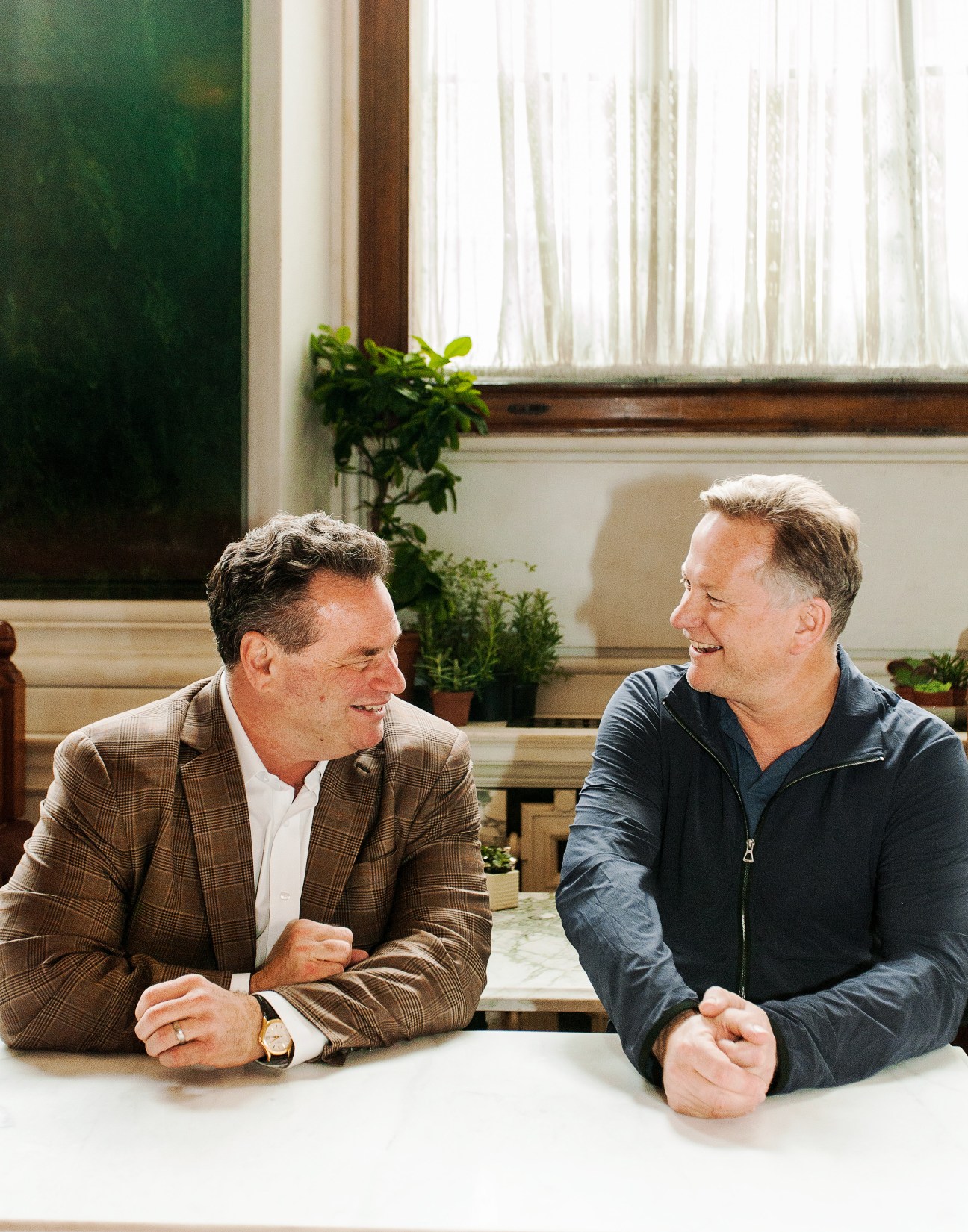 Andrew Zobler and Nick Jones seated at a marble counter in The Ned, conversing and smiling.