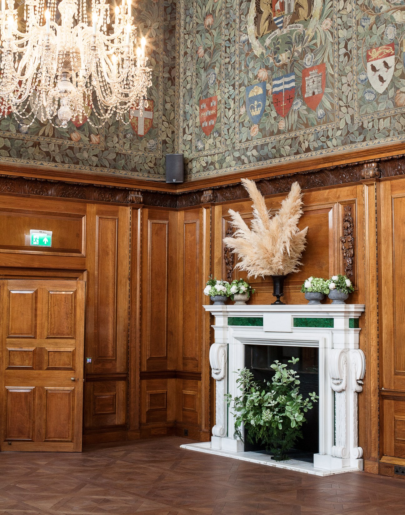 An ornate room with panelled wood walls, decorative tapestry, crystal chandelier and white fireplace mantel at The Ned.