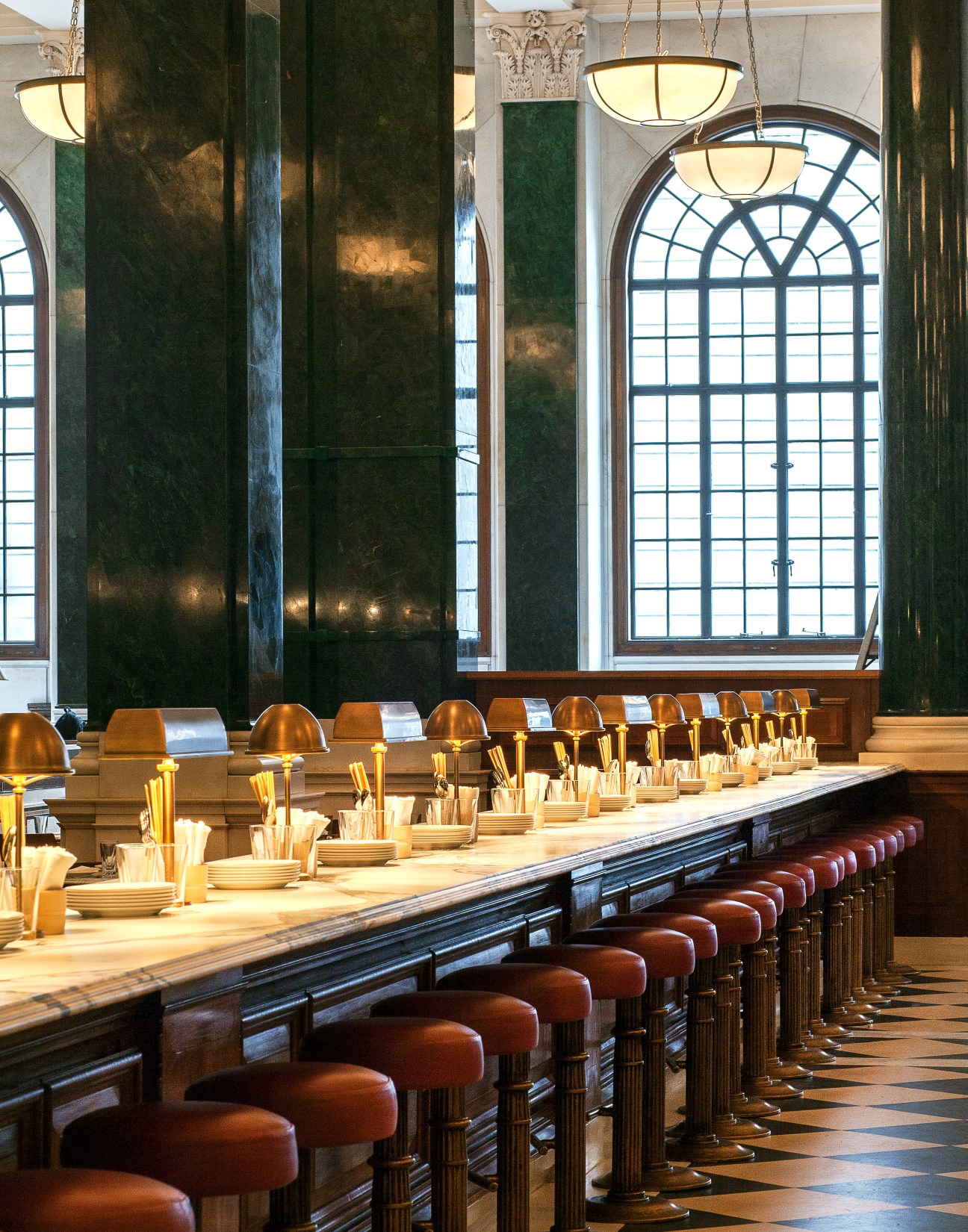 Long dining counter with brass cloche-covered place settings beneath soaring green marble columns and arched windows in The Ned.