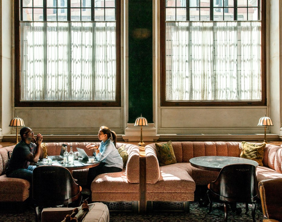 Two people seated at a table in The Ned's lounge area with large windows, decorative lamps, and upholstered seating.
