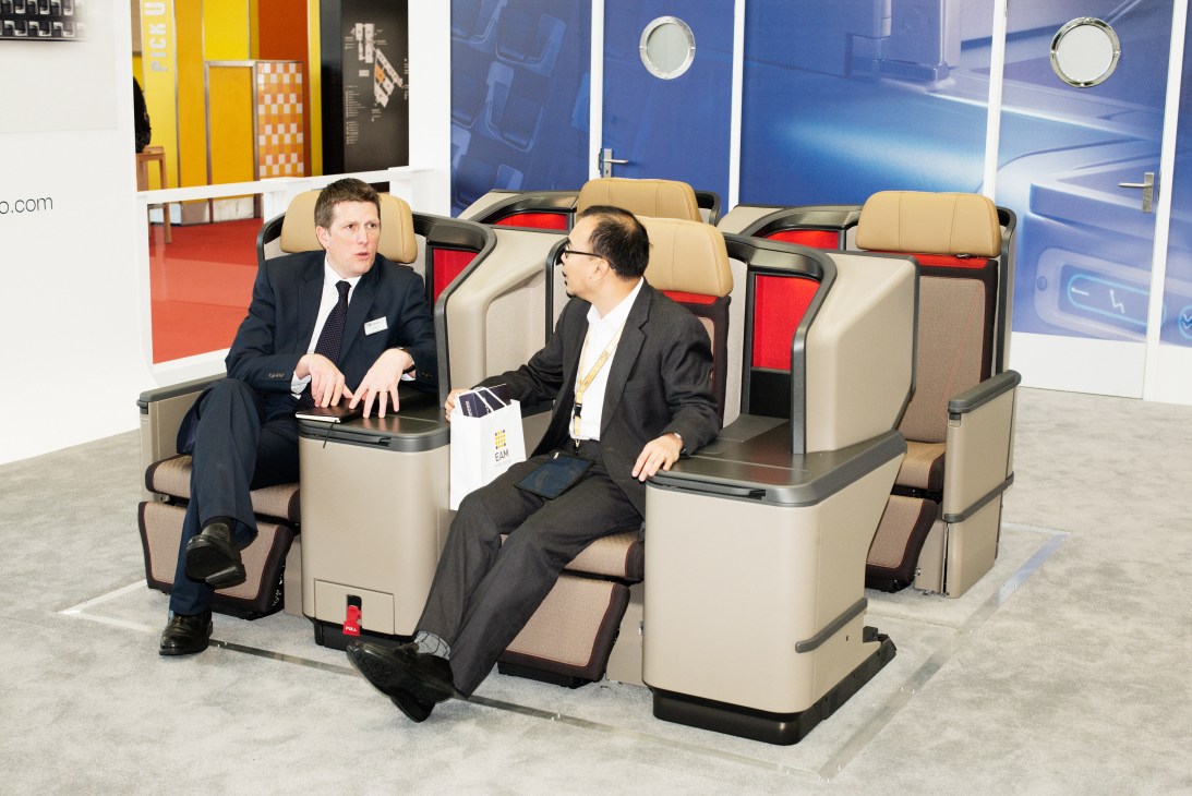Two men in business suits sit in beige and red aircraft business class seats at an expo booth.