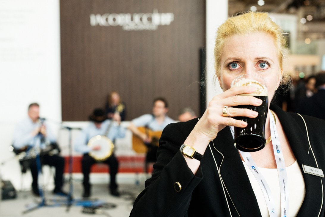 Woman drinking dark beer at Aircraft Interiors Expo with Irish band performing in background.