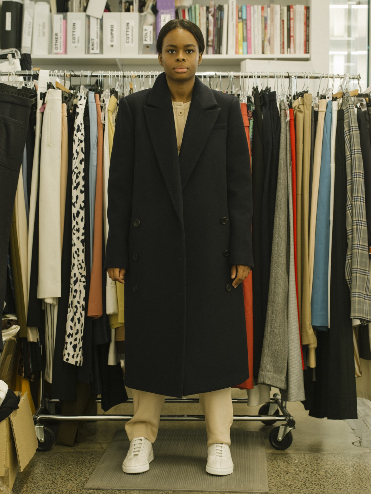 A woman wearing a navy overcoat and white trainers stands in front of clothing racks in a studio.