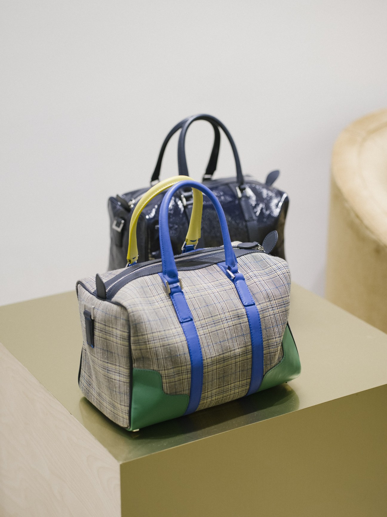Two Tibi handbags stacked together: a checked fabric tote with blue and yellow straps and a navy leather bag behind it.