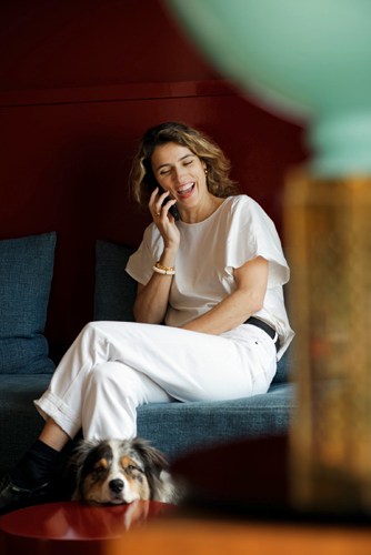 Woman in white clothing sitting on a blue couch while on a phone call, with a dog at her feet.