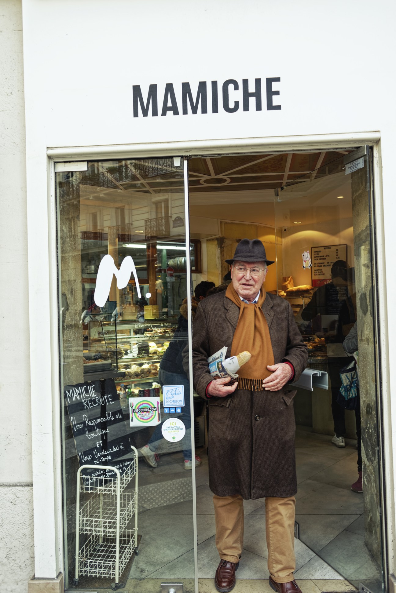 Man in brown coat and hat stands outside Mamiche bakery holding bread product.
