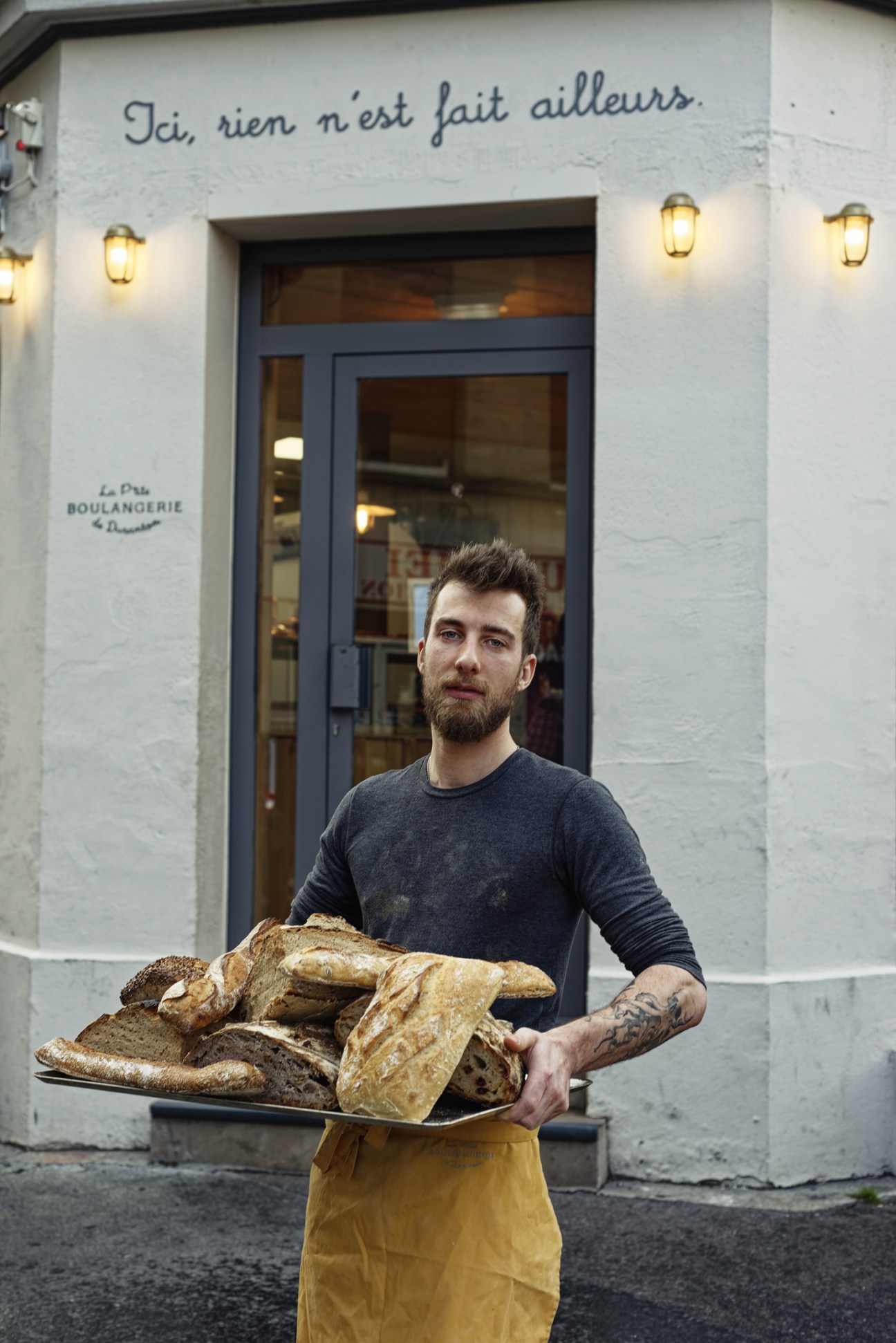 A bearded man in a dark sweater holds a tray of freshly baked bread outside P'tite Boulangerie.