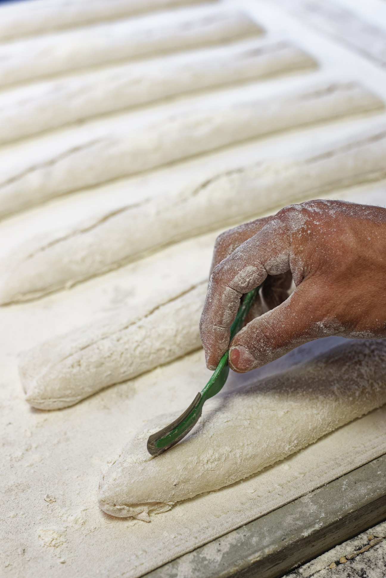 A baker's flour-dusted hand shapes dough with a green scraper tool on a work surface lined with white dough.