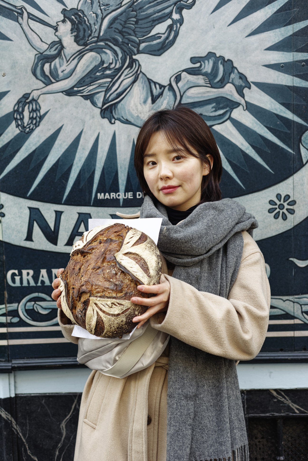 Woman holding a round artisanal boule loaf of bread with scored leaf pattern in front of decorative mural.