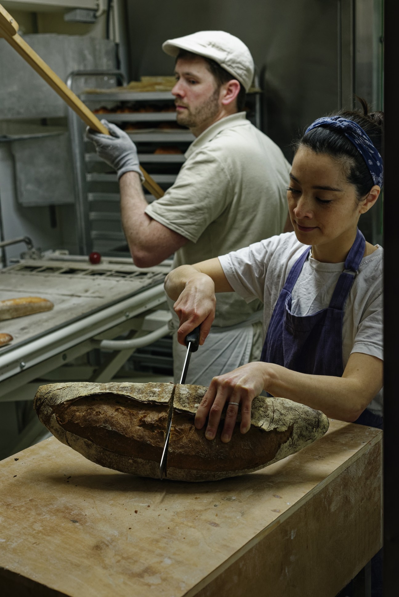 Female baker scoring a round loaf of bread with a scoring tool in an artisanal bakery kitchen.