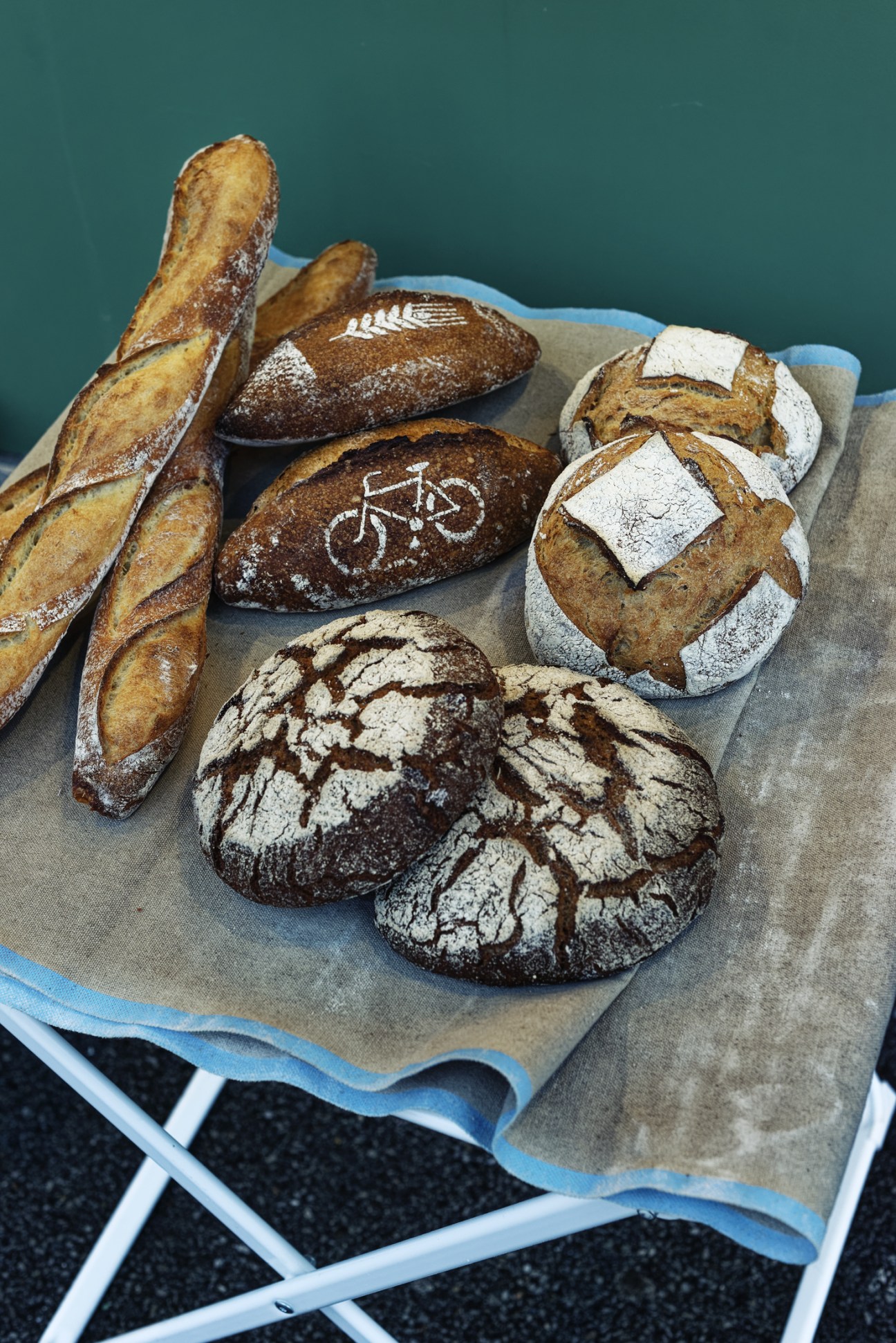 Assorted artisanal breads including baguettes, boules, and round loaves dusted with flour on parchment paper against a teal background.