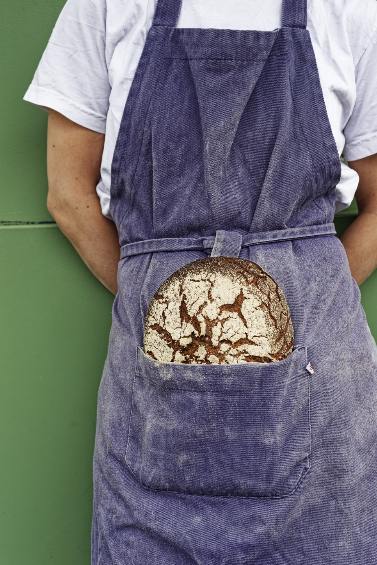 Baker in denim apron holding a crusty round loaf of bread against green background.