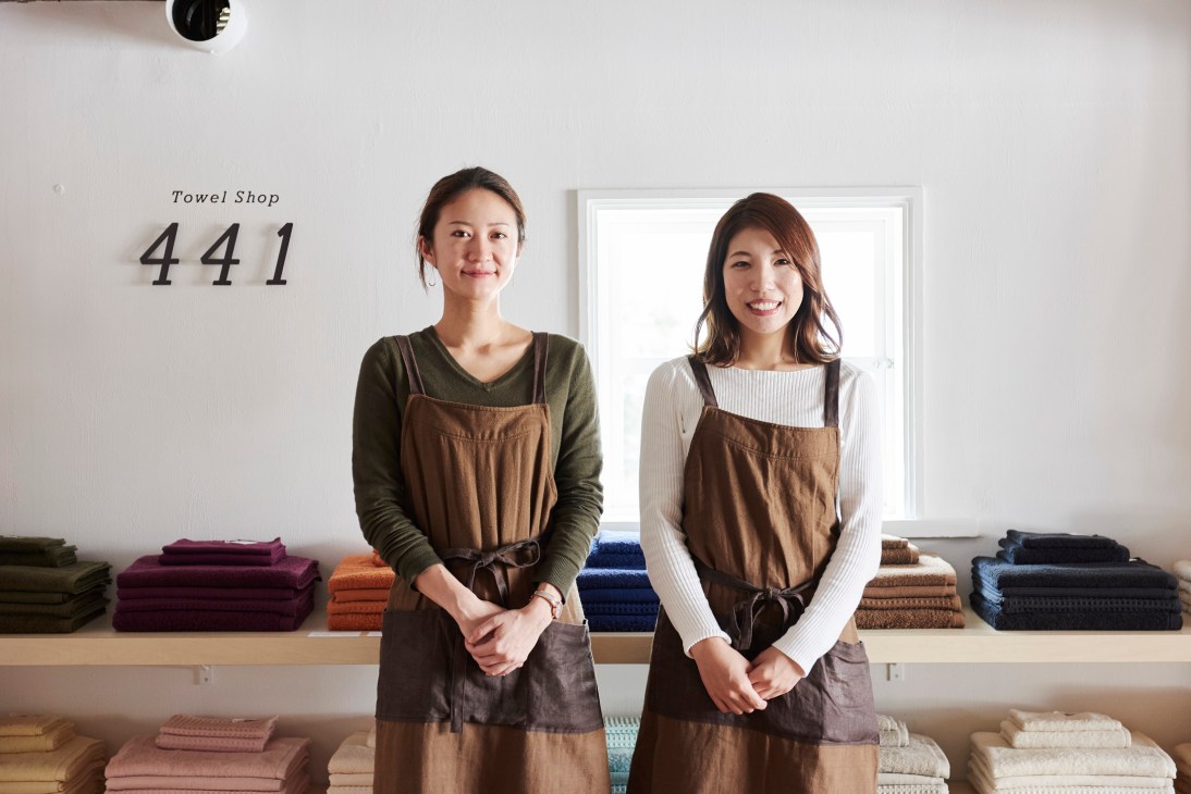 Two female staff members in brown aprons standing in front of stacked towels at Towel Shop 441 in Tokyo.
