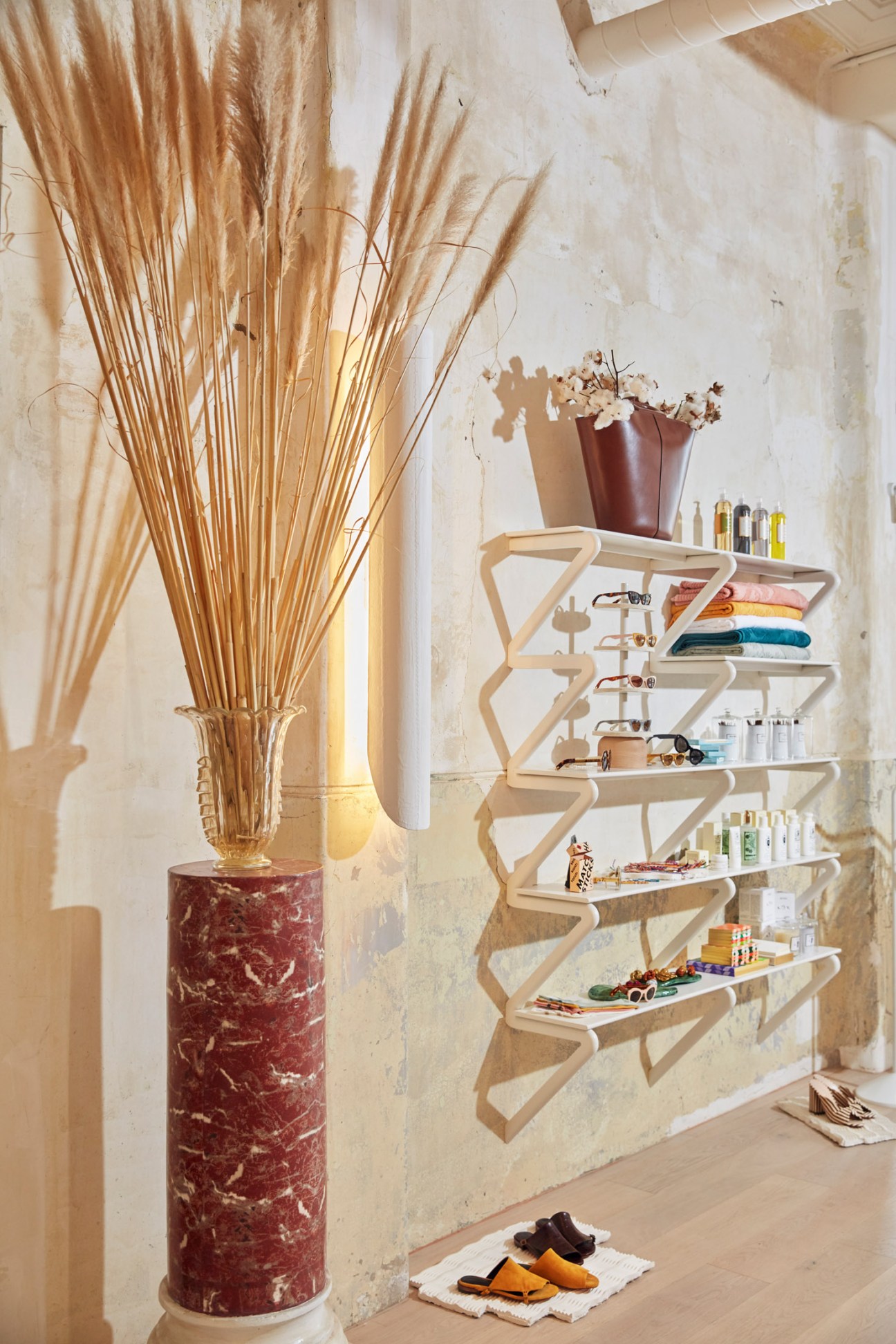 Sculptural zigzag shelving unit displaying colorful products next to dried wheat stalks in red marble vase