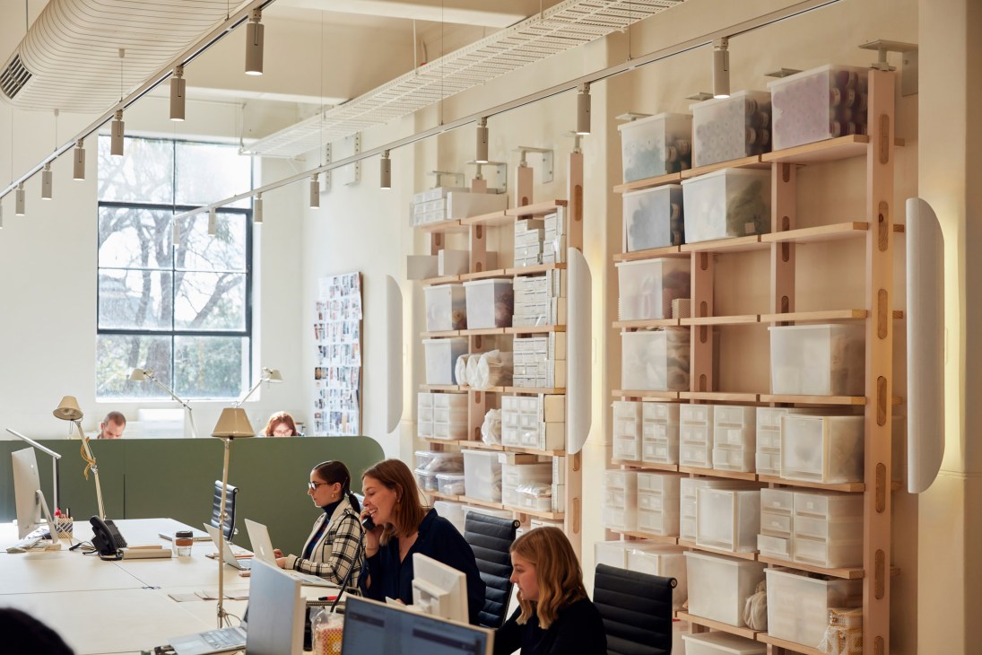 Modern office workspace with wooden shelving units displaying white storage boxes and design samples, two women working at desks in foreground with large windows overlooking trees.